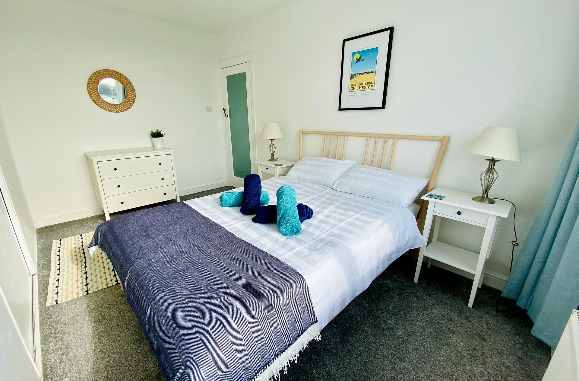 Ocean Grove Image A large double bed sits at the centre of a bedroom styled in greens and blues. A set of blue towels are rolled up on the centre of the bed and a circular mirror hangs on the wall.