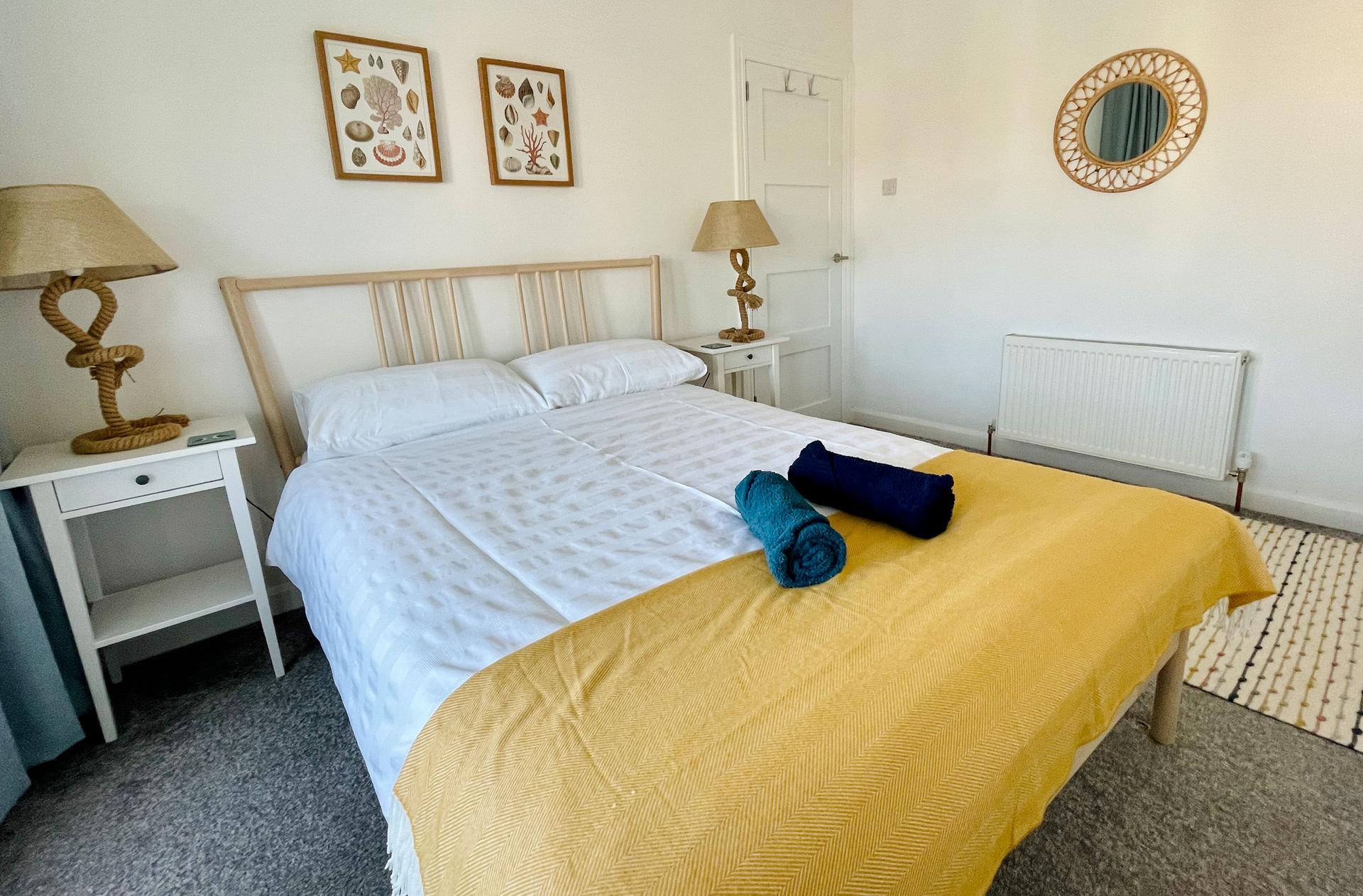 Ocean Grove Image A large double bed in the centre of a room styled in yellows. A set of towels are rolled up on the bed and a circular mirror hangs on the wall.