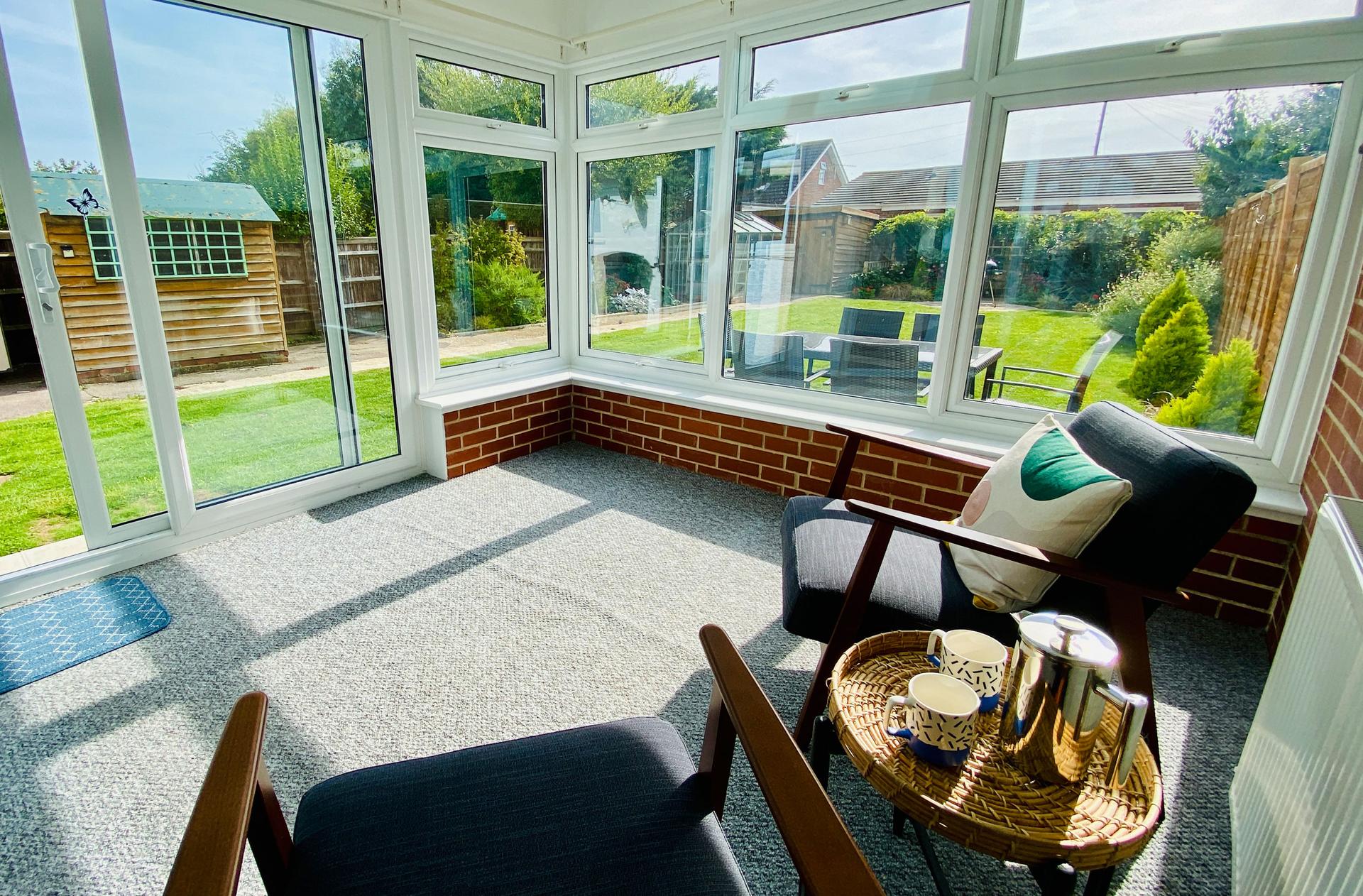 Ocean Grove Image A conservatory seating area looks out towards a large spacious garden with a set of table and chairs and a barbeque in the background.