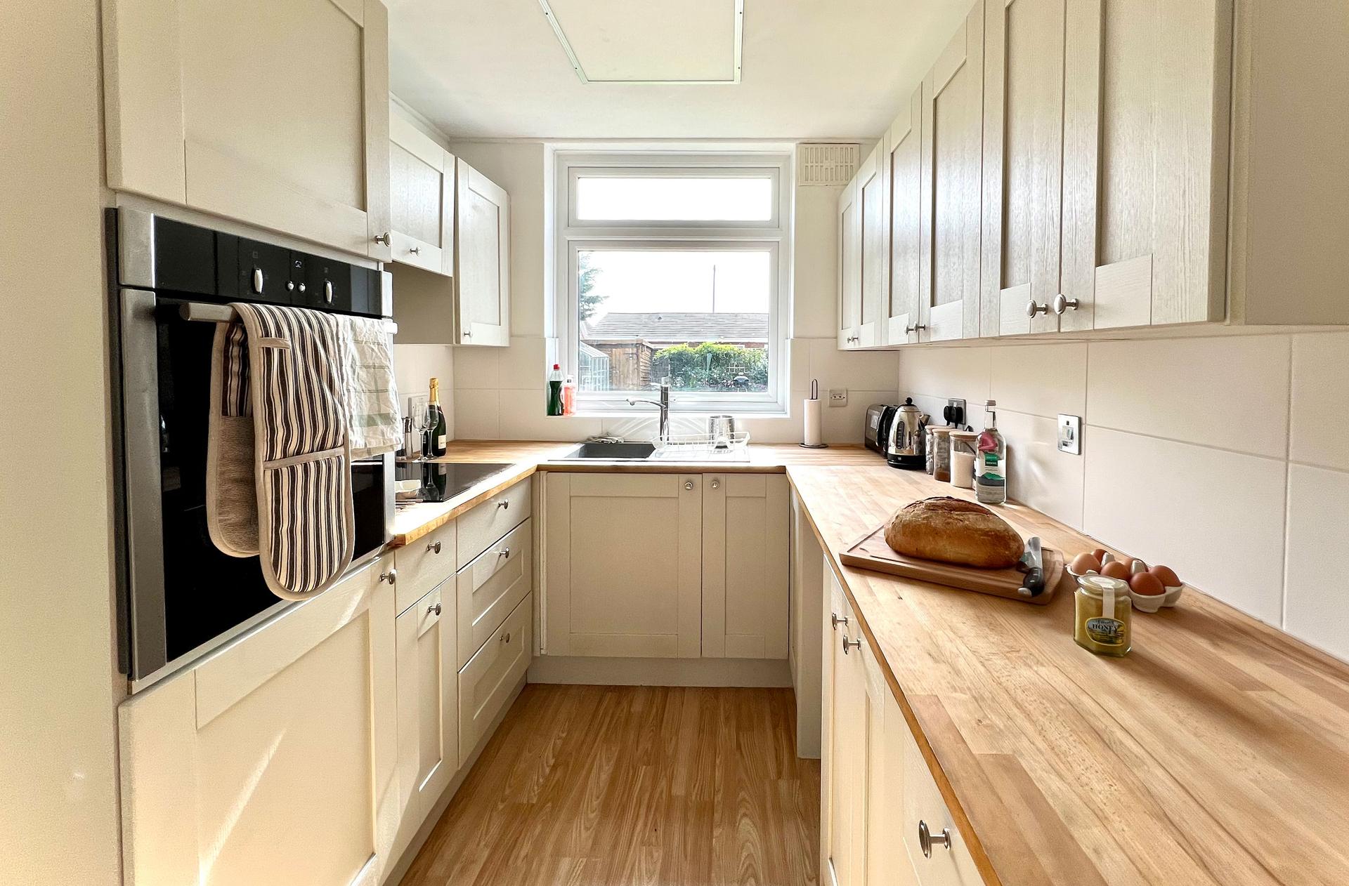 Ocean Grove Image A small kitchen with clean work surfaces featuring a loaf of bread and a glove mitt hanging on the oven on the left. A window looks out onto the view outside.