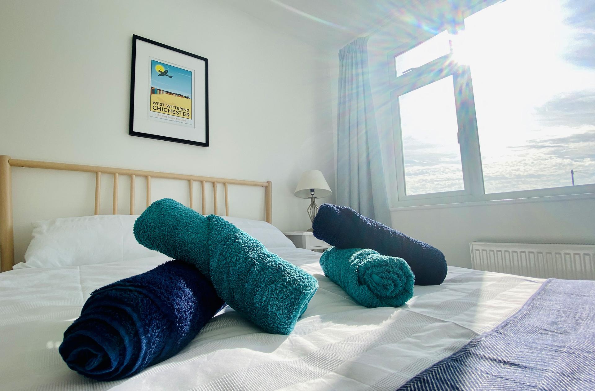 Ocean Grove Image A close up set of towels rolled up on a bed with a framed poster of the West Witterings Chichester. Light pours into the room on a bright day.