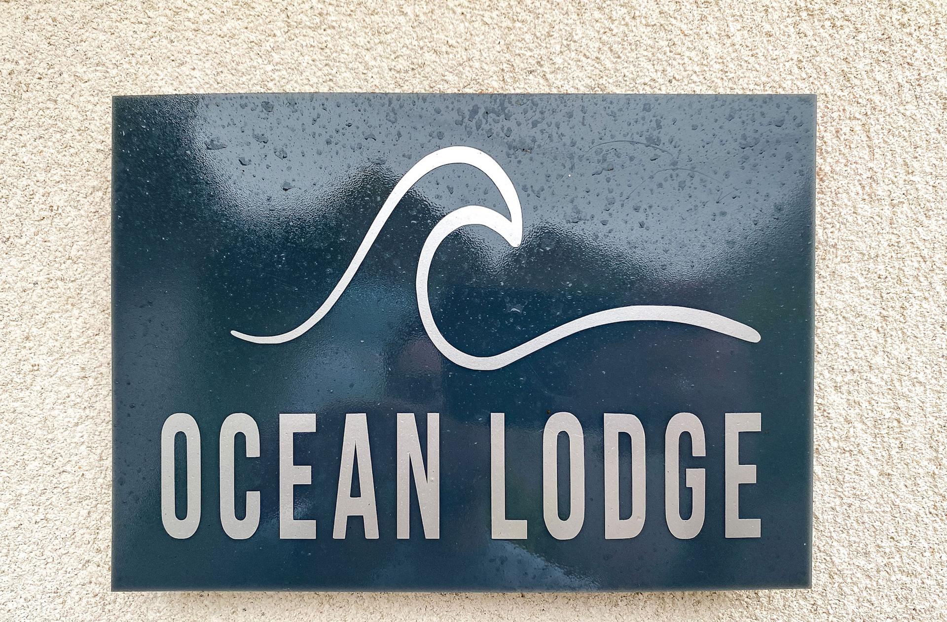 Ocean Lodge Image The words Ocean Lodge and a wave are written on a blue plaque hanging on a sandy coloured wall.