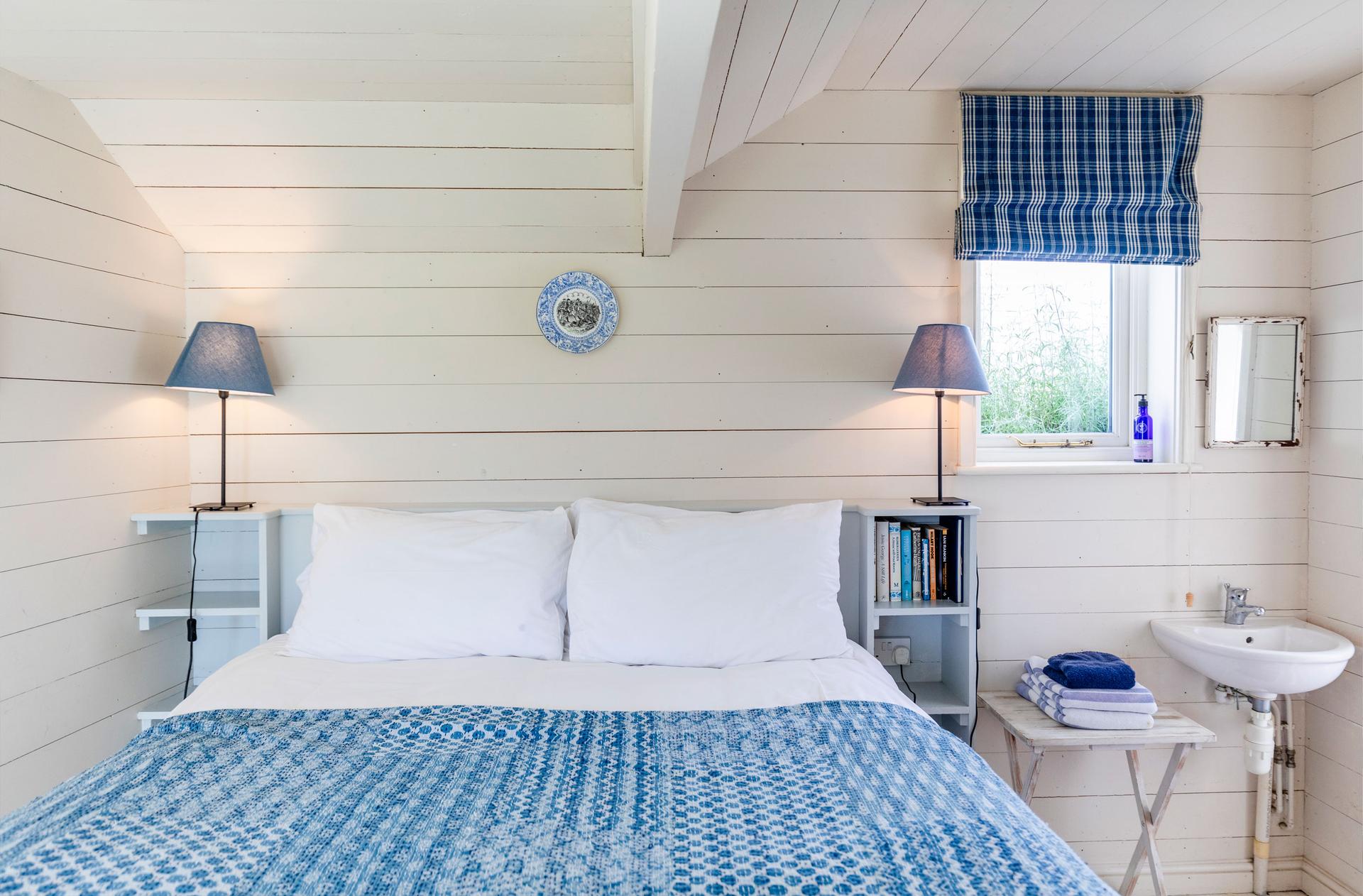 The Dodo Image A blue double bed with a set of white pillows and two lamps placed on the left of an open window with a small sink beneath and to the right.